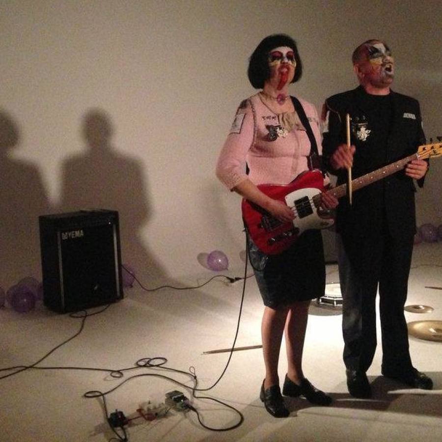 Photo of Chrissie Butler and mr sterile standing side by side mid performance. Chrissie is wearing a red bass and is playing it, mr sterile is also playing the bass with one hand and a drumstick. No drum kit had been arranged for this show - it was a challenge. Scattered at their feet are gallons, cymbals and a snare drum - all of which had been scattered about in order to make sound.
The show was in a small art venue in Beijing, China.