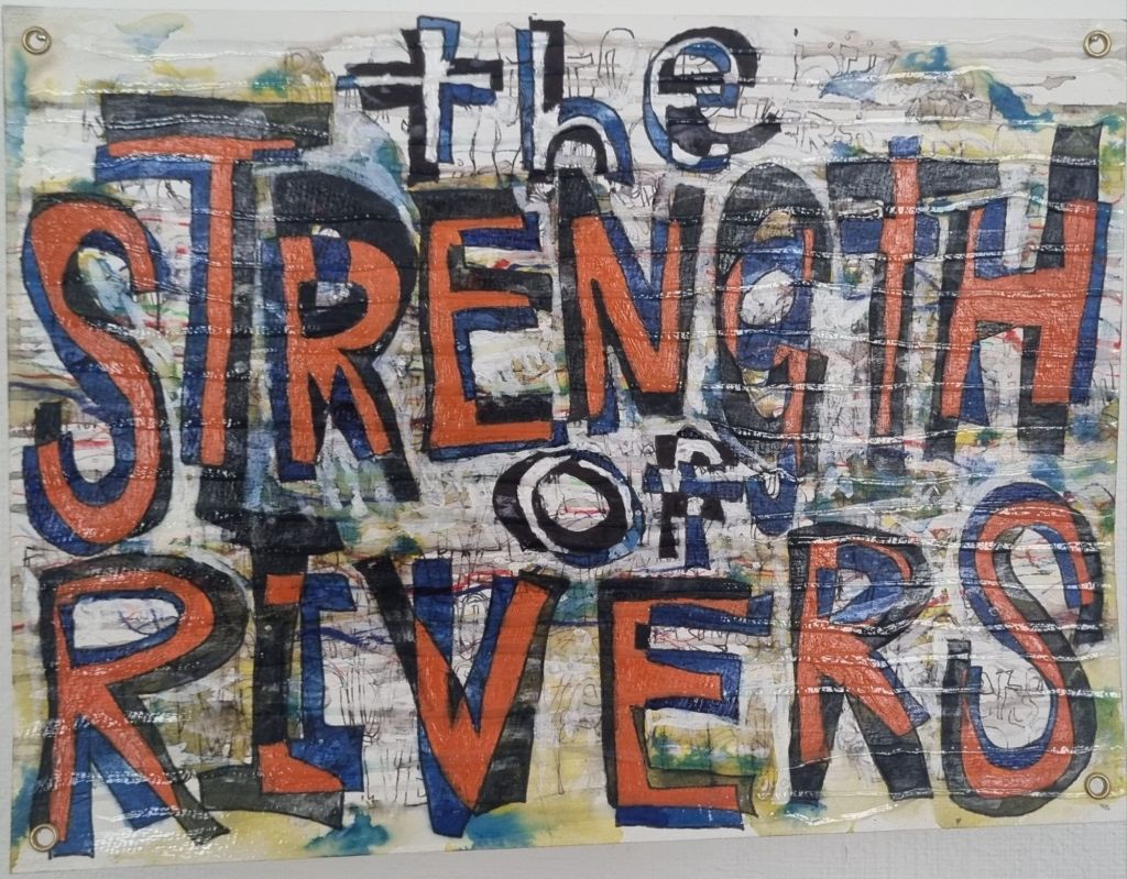 A picture by Kieran. It says:
the Strength of Rivers.
The words Strength and Rivers are uppercase, in black, orange and blue.
The words the and of, are lower case in blue, black and white.
The background is scratchy, drawn on, and covered-over streaks of ink, splashes of yellow and blue watercolour.